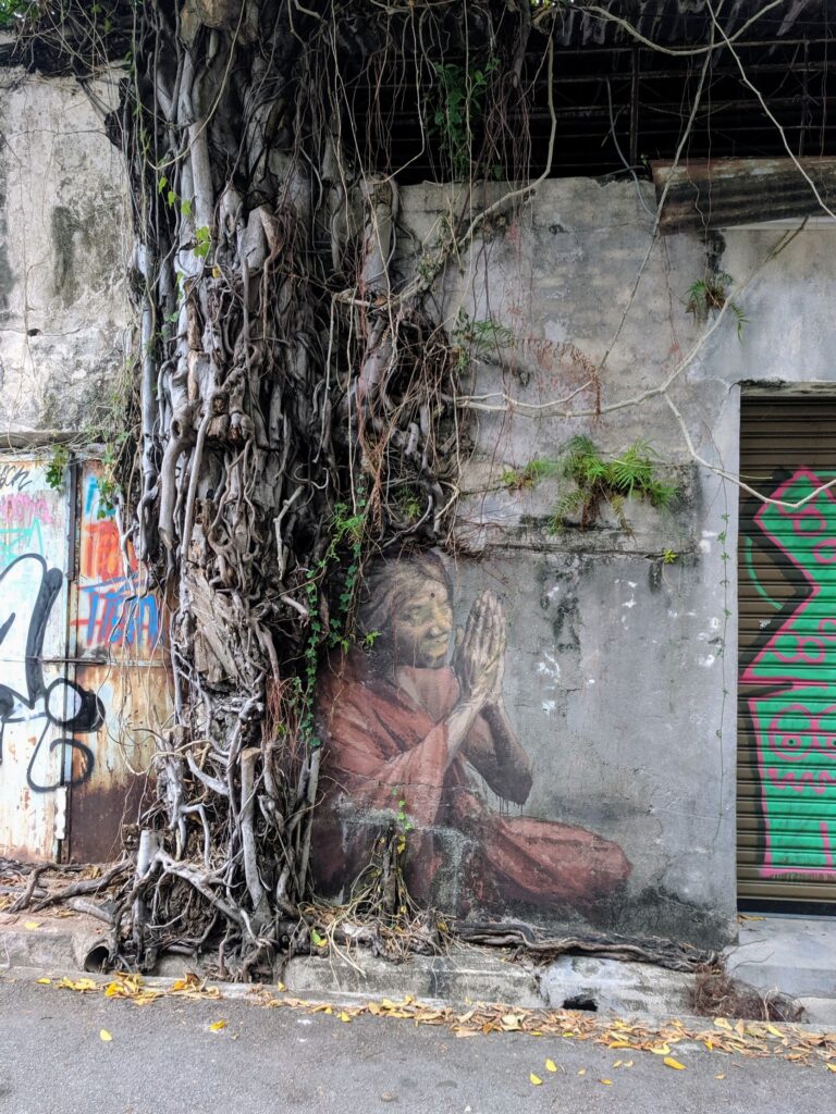 An old tree growing up a wall with a painting of a woman sitting on its roots