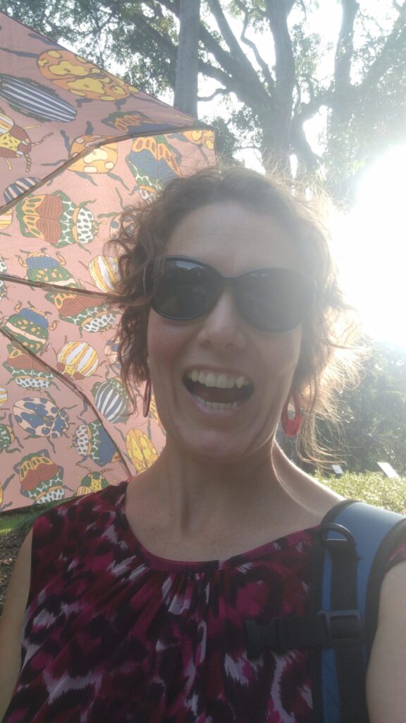A woman smiling at the camera holding an umbrella with the sun poking through the trees behind