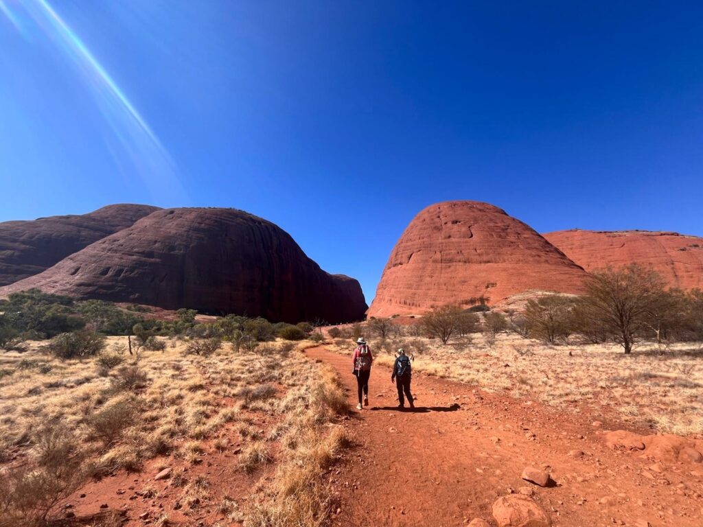 Two women walking on a red dirt path towards two large rocks with a gorge inbetween