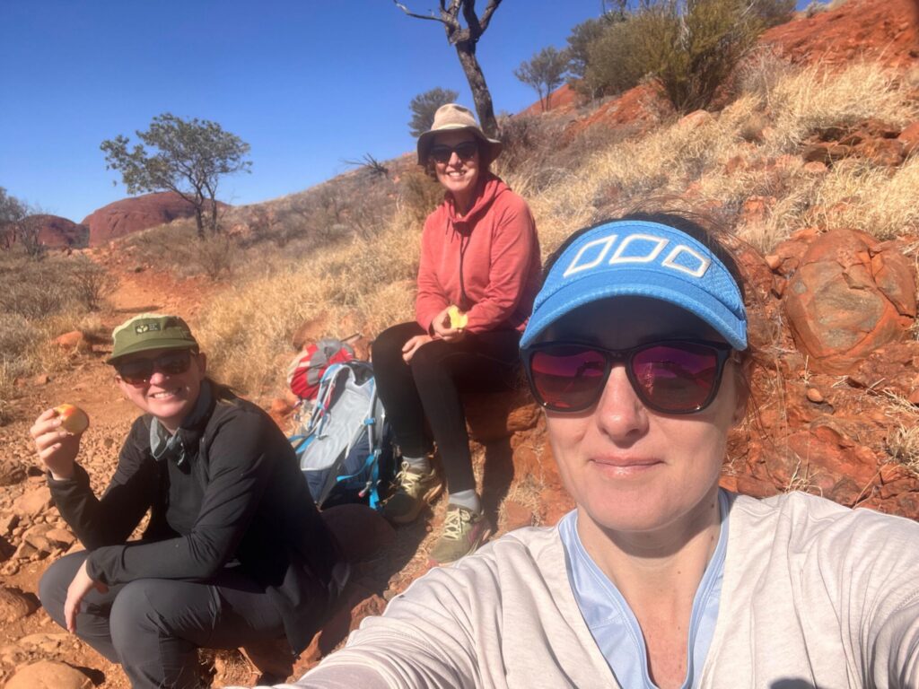 A selfie of three hikers, sitting eating apples in a red desert landscape