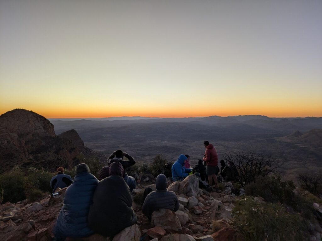 A handful of people sitting on top of a mountain looking into the distance as the sun slowly rises across a dry desert landscape