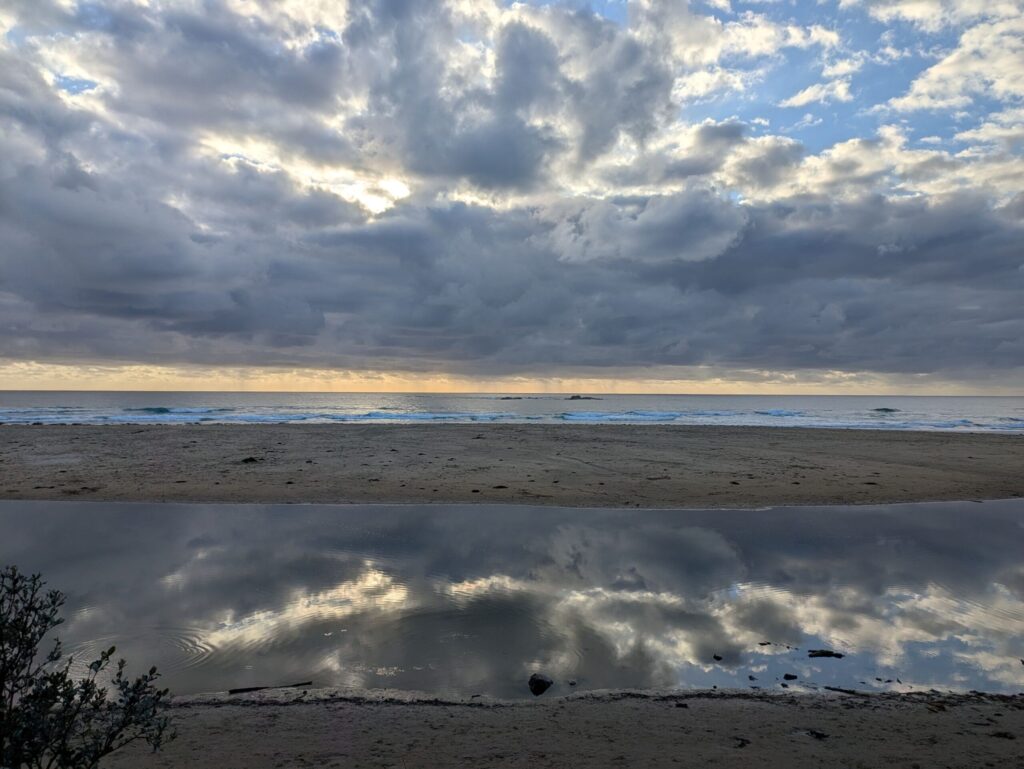 Looking out to the ocean from a beach, with a reflection of the clouds in a lagoon in the foreground