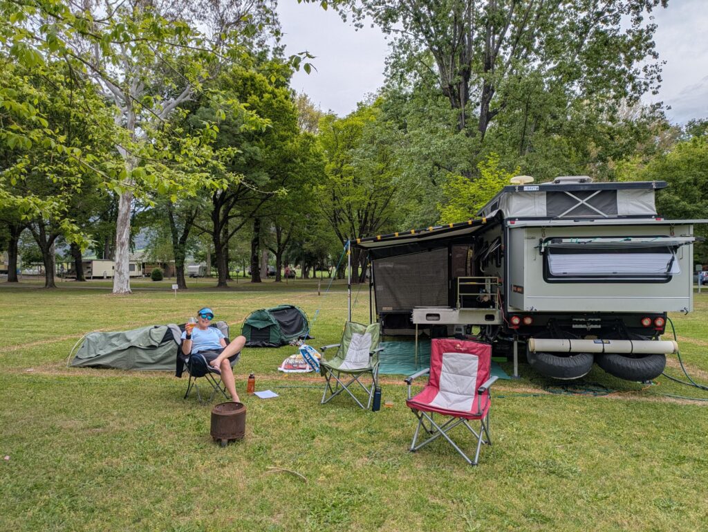 A woman sitting outside a caravan with swags nearby, 2 chairs for friends in a lush green campground