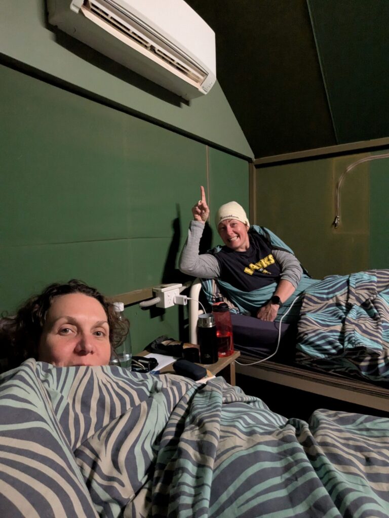 Two women in single beds in a tent, pointing to the air conditioner attached above them