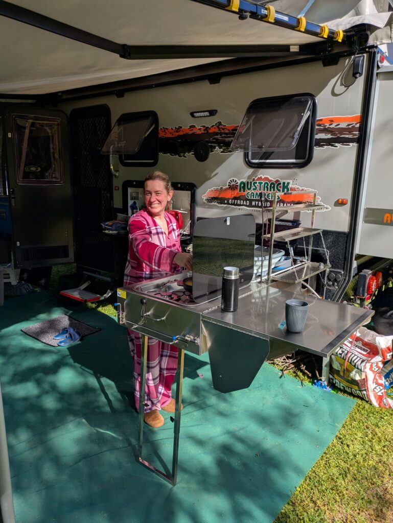 A woman smiling at the camera while she cooks breakfast on a stove attached to a caravan