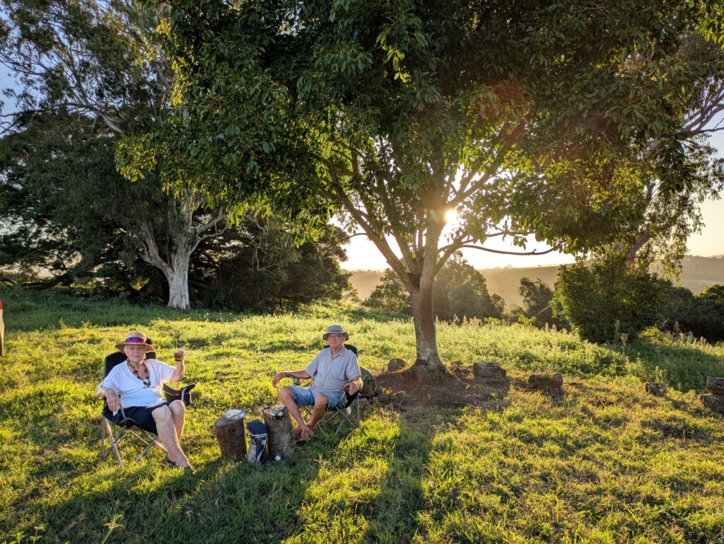 two friends sitting by a tree with a small picnic and the sun setting behind them