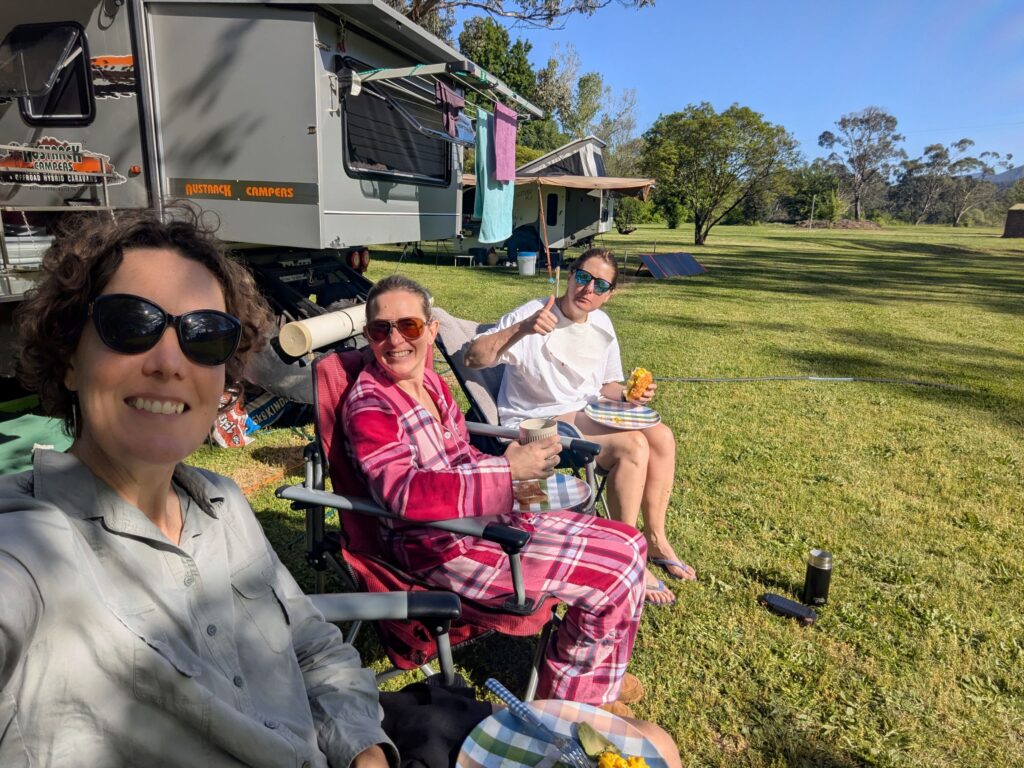 Three friends sitting in camp chairs eating breakfast in the morning sunshine