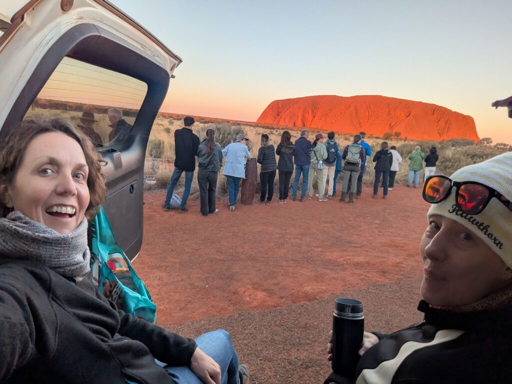 Two friends sitting in a car watching a crowd of tourists jostle for a position to watch the sunset at Uluru