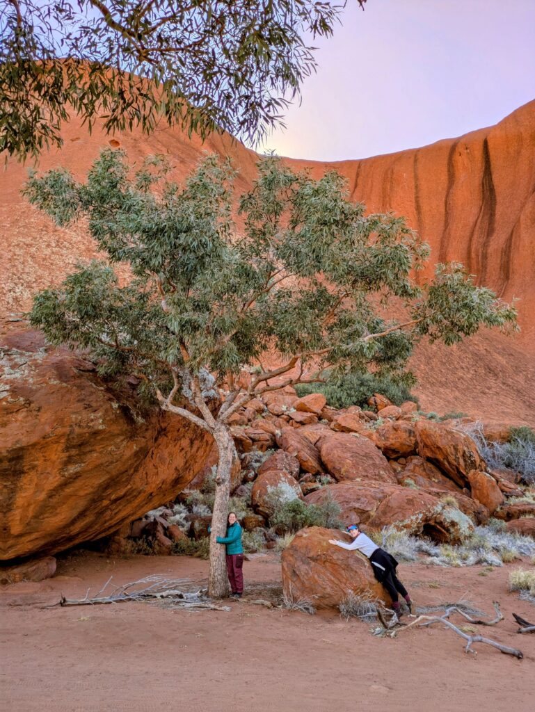 Two women at the base of Uluru - one hugging a tree and one hugging a rock