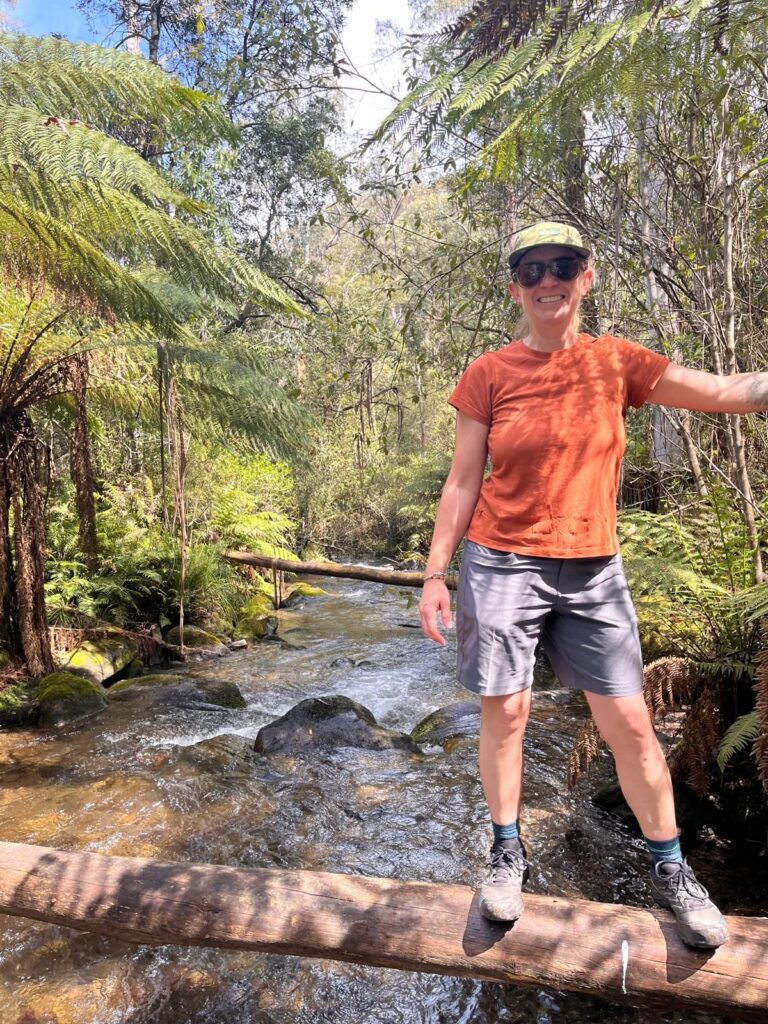 A woman in the foreground standing on a log over a running creek with dense forest behind her