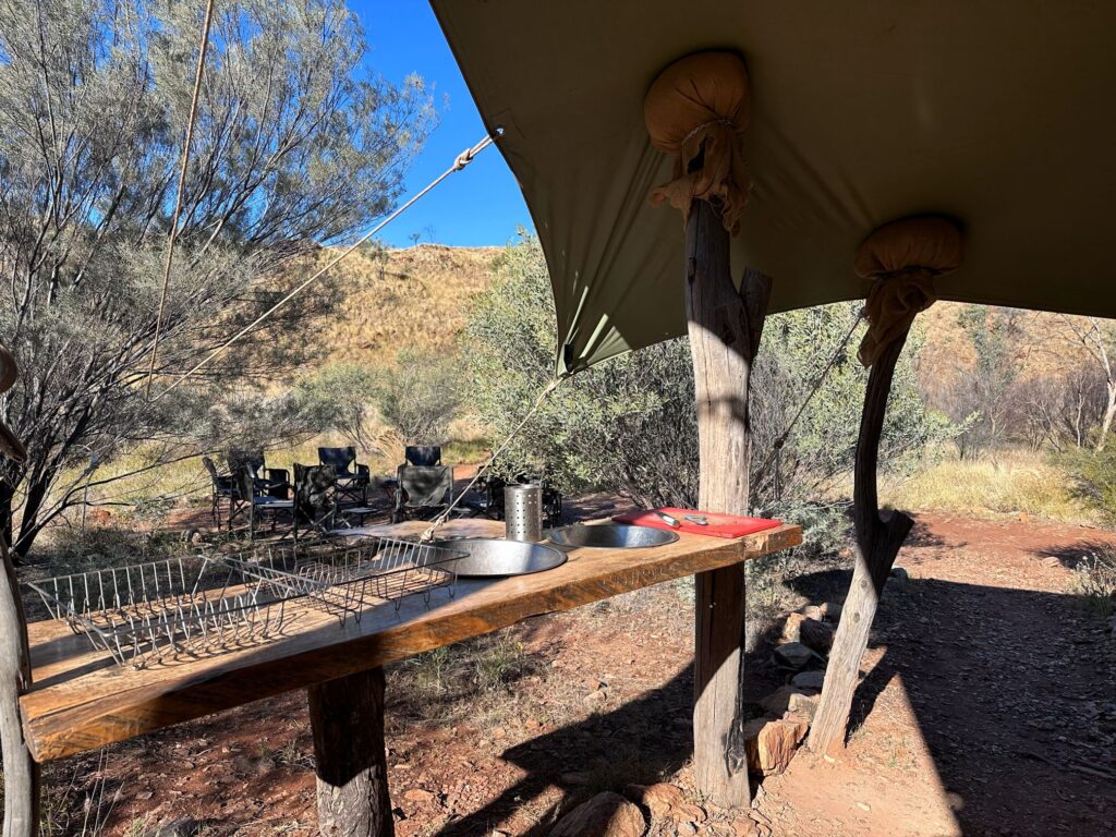 A high bench with two bowls set into for washing up under a tarpaulin structure, part of an outdoor kitchen