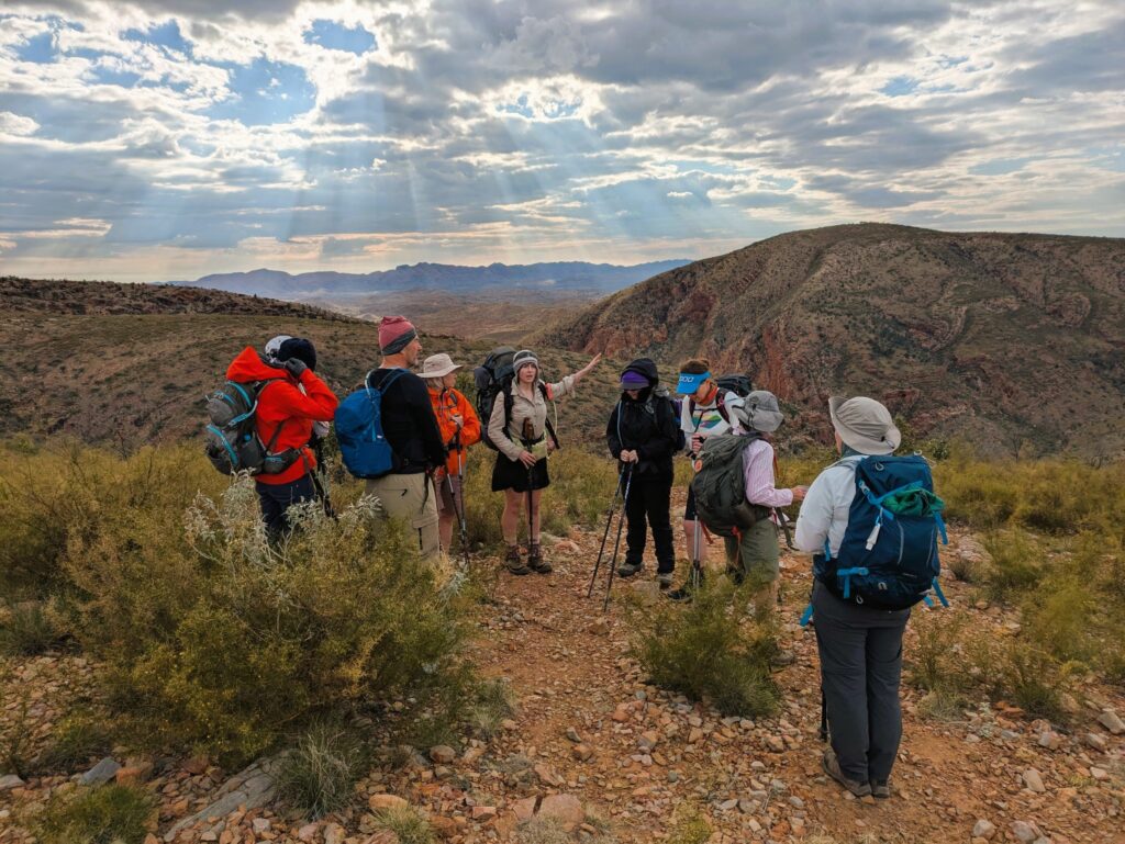 A tour guide telling a group of hikers about the area in which they are walking. Large mountains in the distance and the sun breaking through the clouds