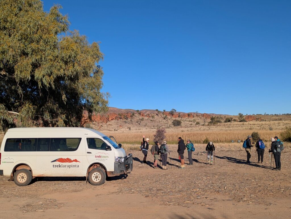 A mini-van parked near a tree on a dry river bed and people standing beside it, getting ready to hike for the day