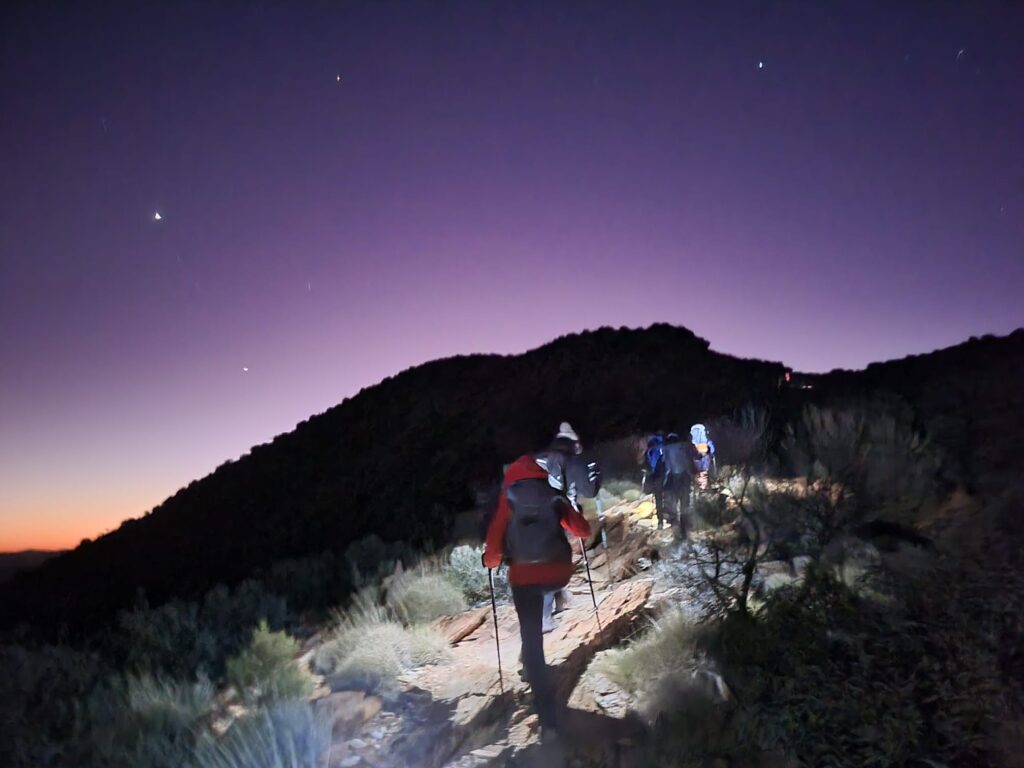 A group of hikers ascending a mountain in the dark, wearing headtorches with a handful of stars still visible as the first signs of daylight appear