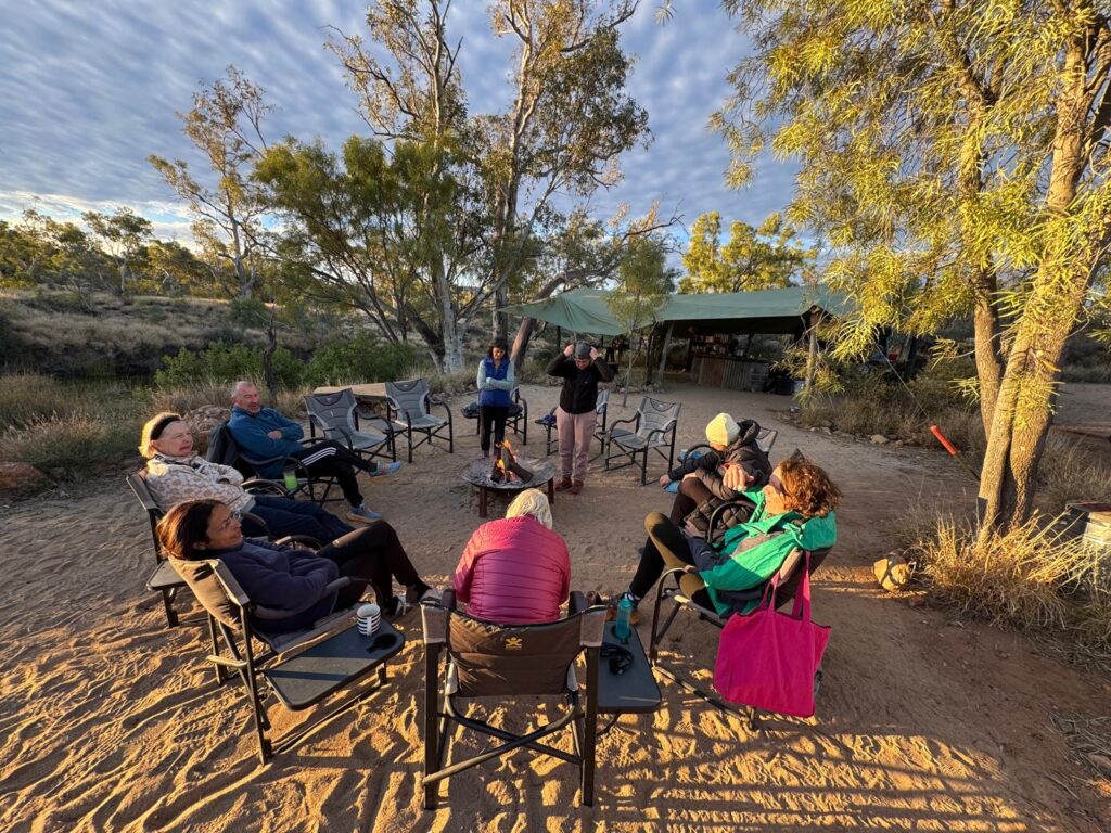 A group of people seated in a circle around a campfire outdoors with long shadows forming as the sun sets
