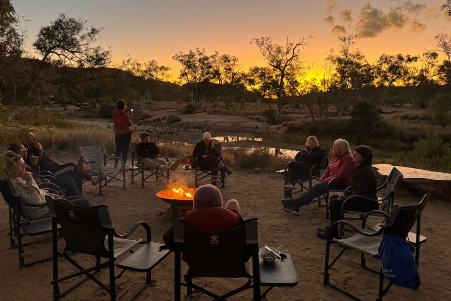 People sitting on chairs in a circle around a firepit in a campground, with a river in the background and the sun setting in the distance