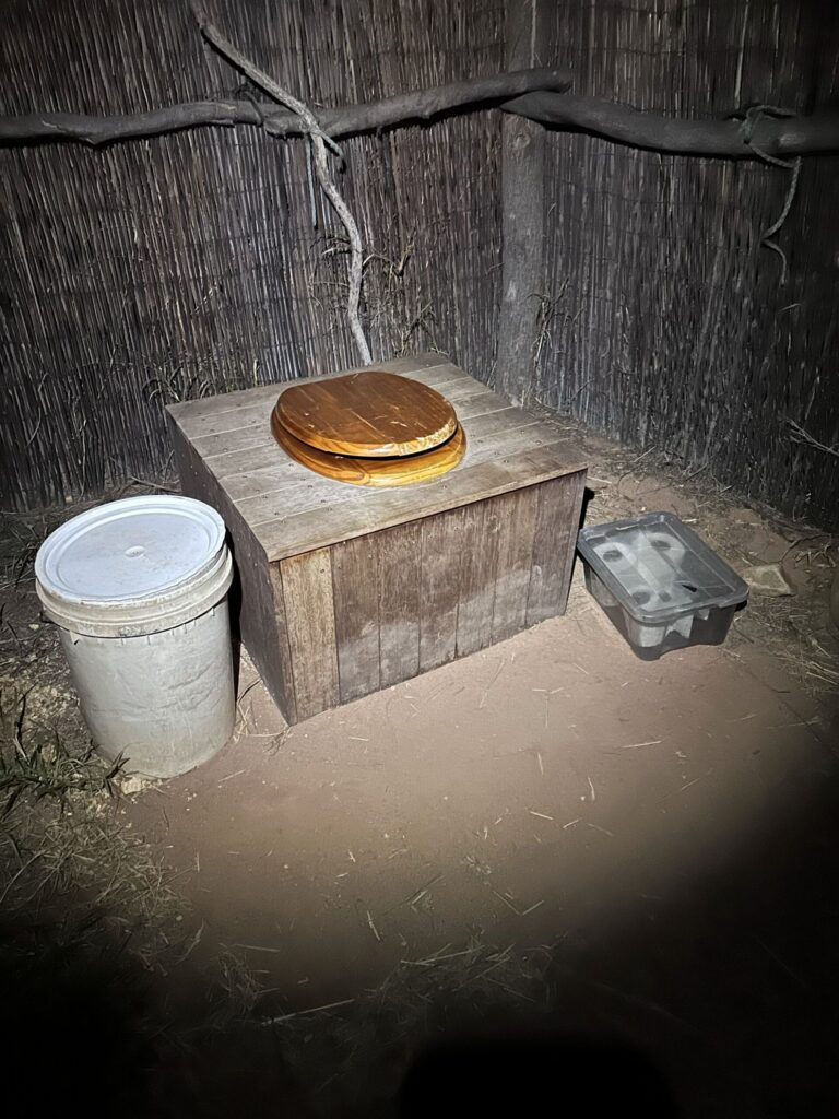 A wooden box with a toilet seat attached, acting as the Eco Loo for a camp. A box of toilet paper sits on one side of the box and a bucket of woodchips on the other side