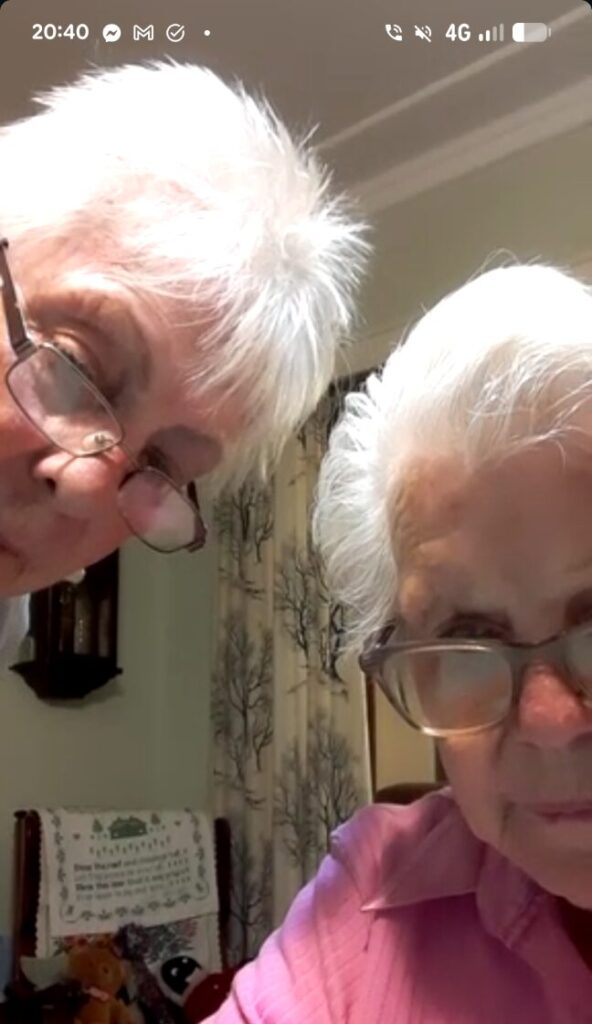 A screenshot on a mobile phone of two elderly women looking at the camera, trying to make the phone work