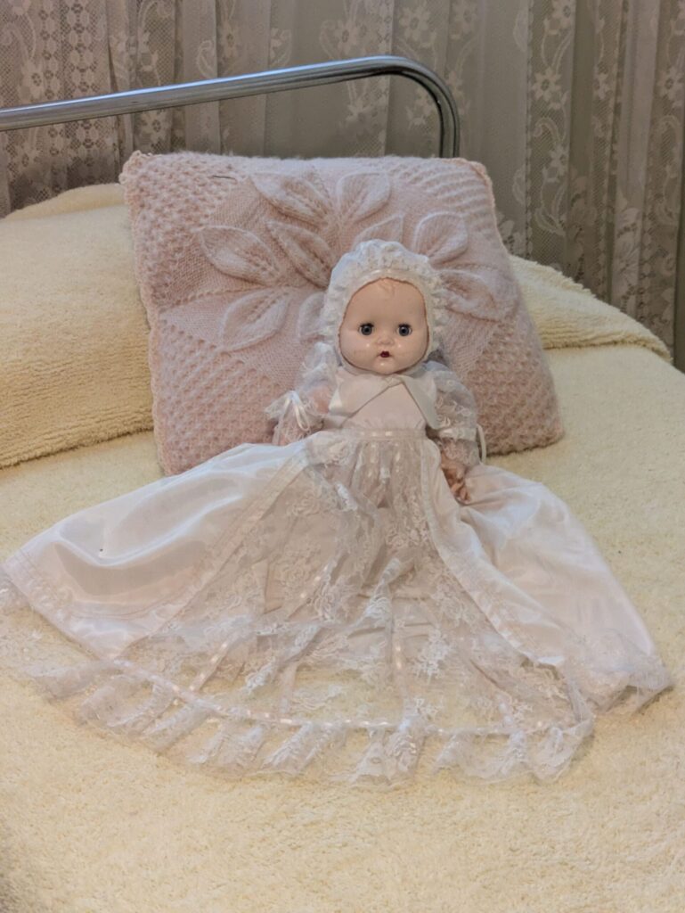 A porcelain doll of a baby in a white lace dress on a bed