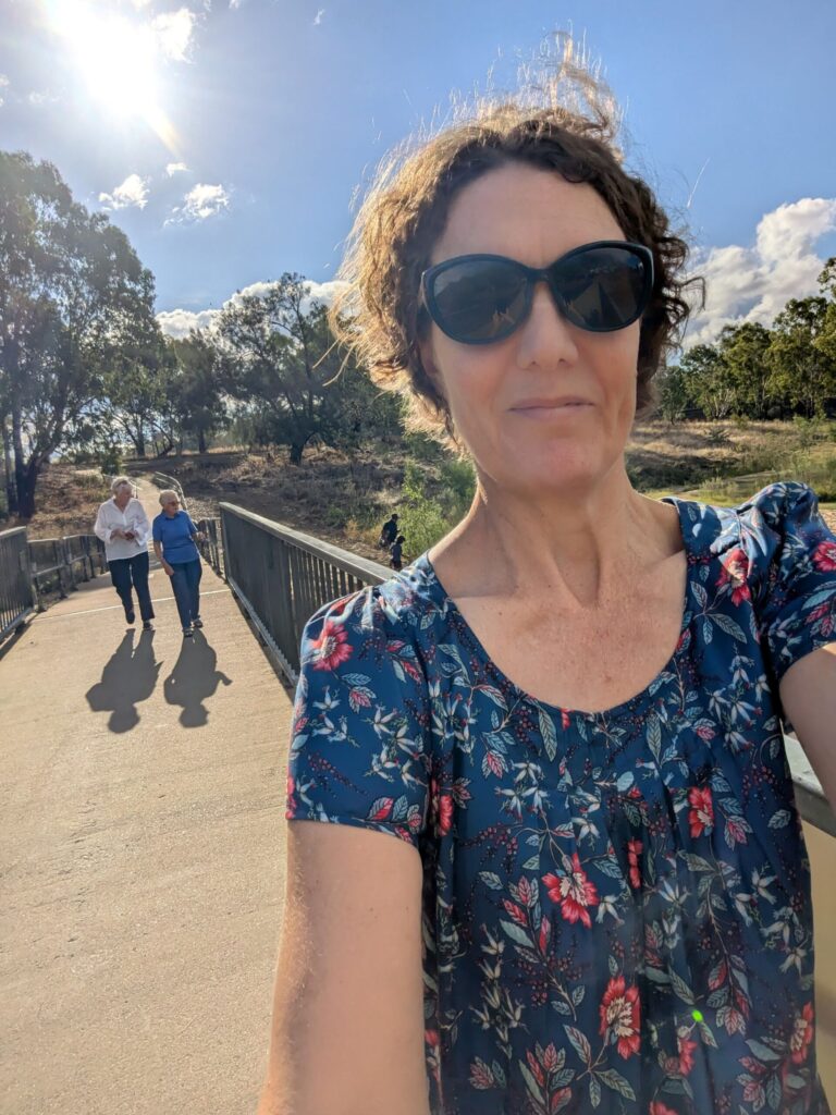A woman taking a selfie with two elderly woman over her shoulder walking along a bridge on a bright sunny day