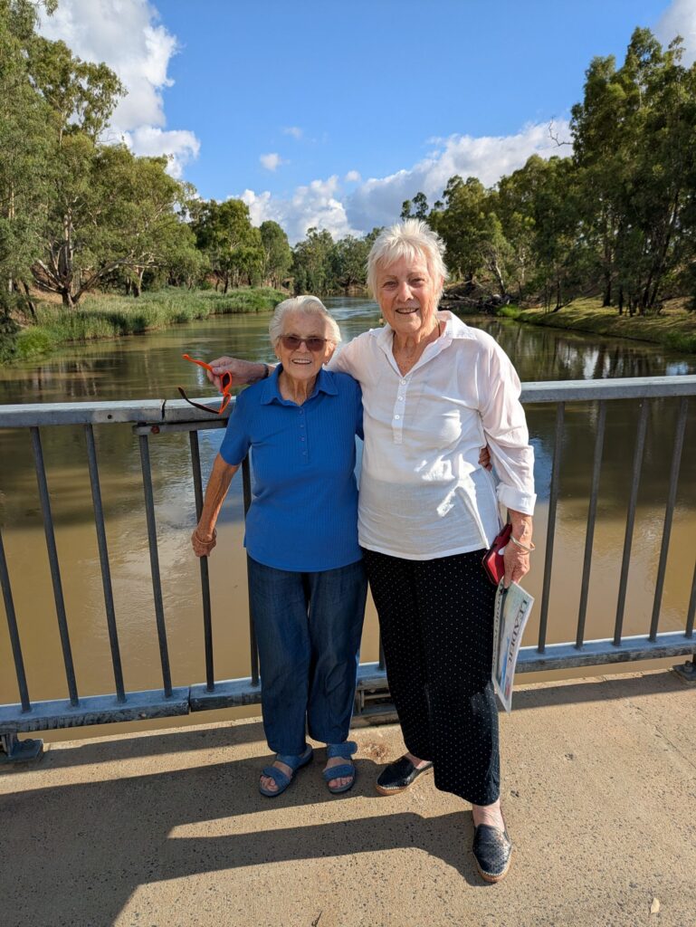 Two elderly women smiling at the camera, standing on a bridge across a river on a bright sunny day