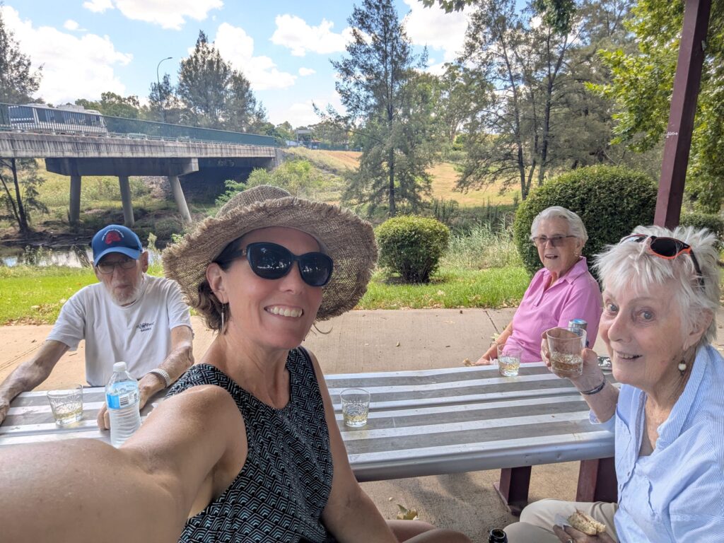 Three woman and an elderly man at a picnic table under a shade cover in a public park enjoying lunch
