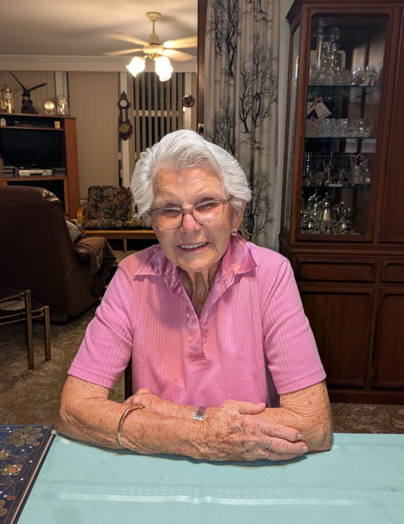 An elderly woman sitting at her dining table in her house, smiling at the camera