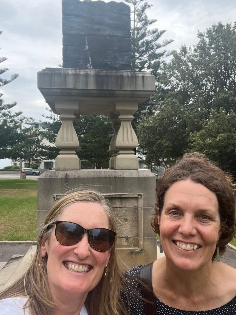 Two women smiling at the camera, standing in front of an outdoor sculpture that appears to be a block of black coal