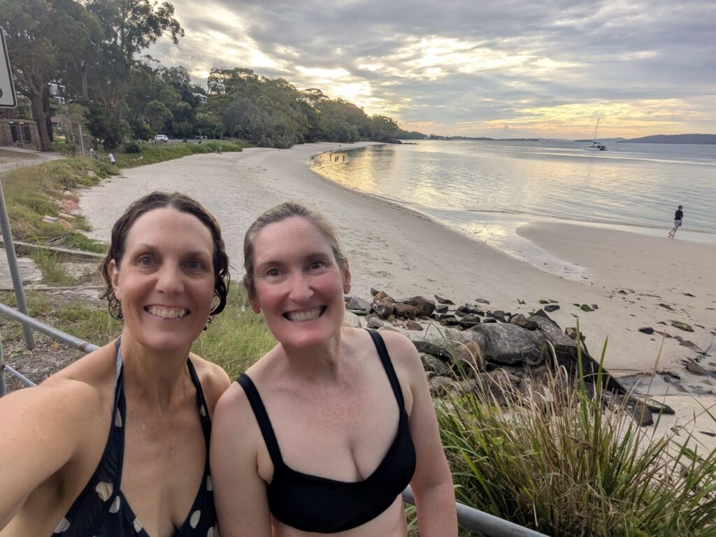 Two women in their swimmers after swimming in a calm bay with a sandy beach which is behind them in the background