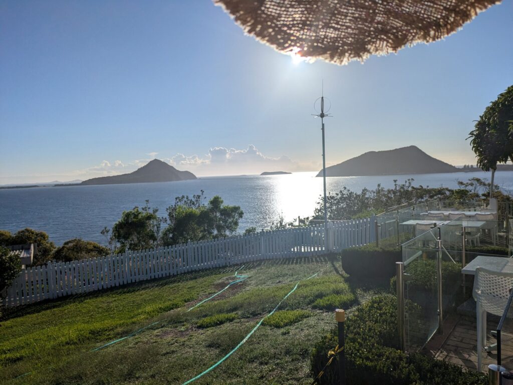 At the base of a lighthouse, looking at the sunrise over the ocean with some small islands in the distance, with a white picket fence and garden furniture on the green lawn