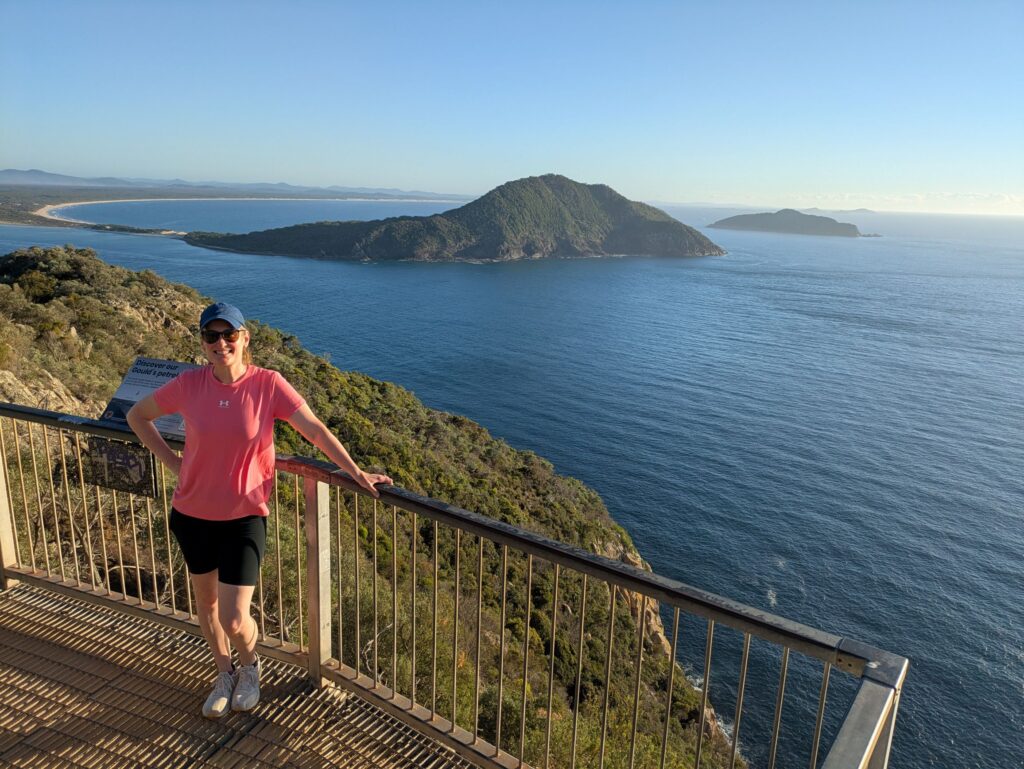 A woman standing at a lookout at the top of a mountain at sunrise with the ocean and a headland behind her