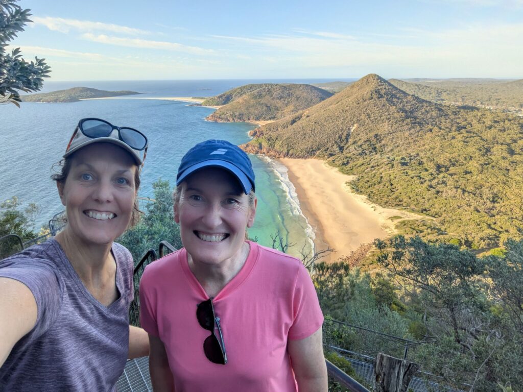 Two friends at a lookout with a series of yellow sandy beaches and small mountains behind them and the big blue ocean