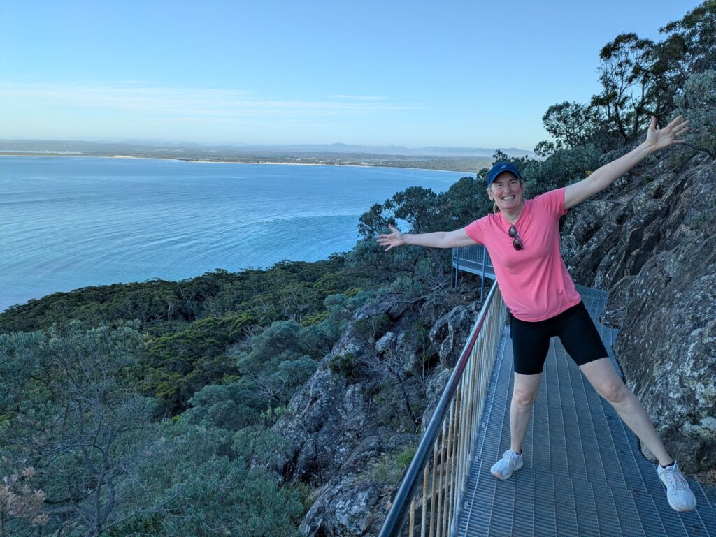 A woman on a raised walkway on the side of a mountain, with her arms and legs out like a star, smiling at the camera, with a river and mountains in the distance.