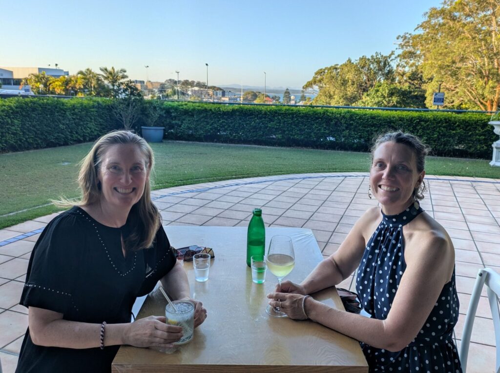 Two female friends sitting at a dinner table outdoors at a restaurant with a view of a town, ocean and mountains in the distance