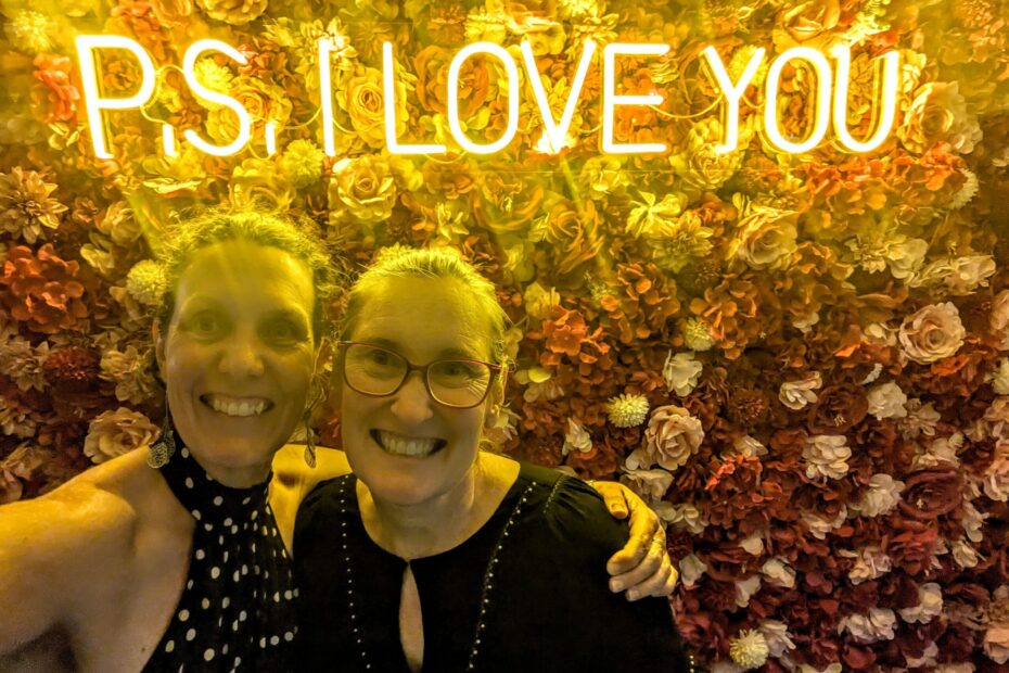 Two friends posing in front of a wall full of flowers with a lighted sign saying "P.S. I love you"