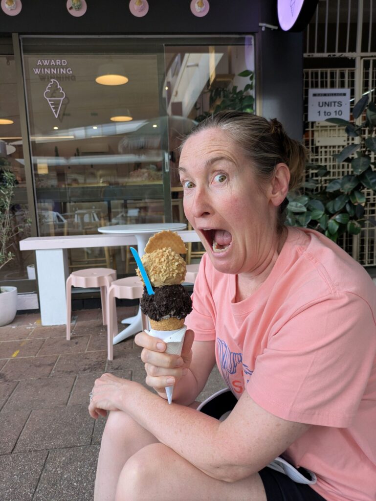 A woman holding an ice cream cone with two large scoops of ice cream, looking at the camera with her mouth wide open about to attack the ice cream