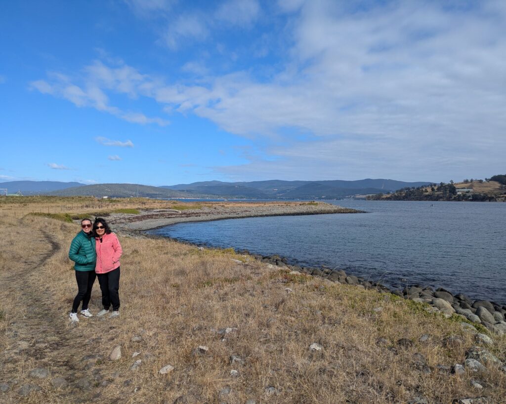 Two friends smiling at the camera at the northern tip of an island with a rocky beach nearby and rolling hills in the distance