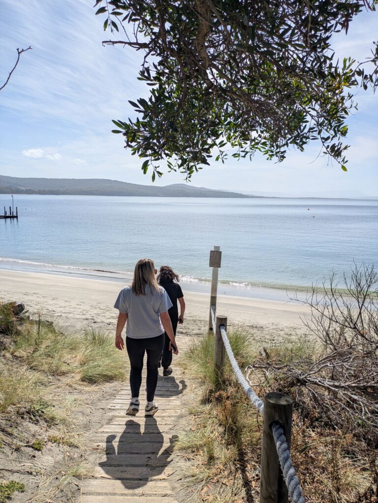 Two friends following a forest path down to a white sandy beach on a sunny day