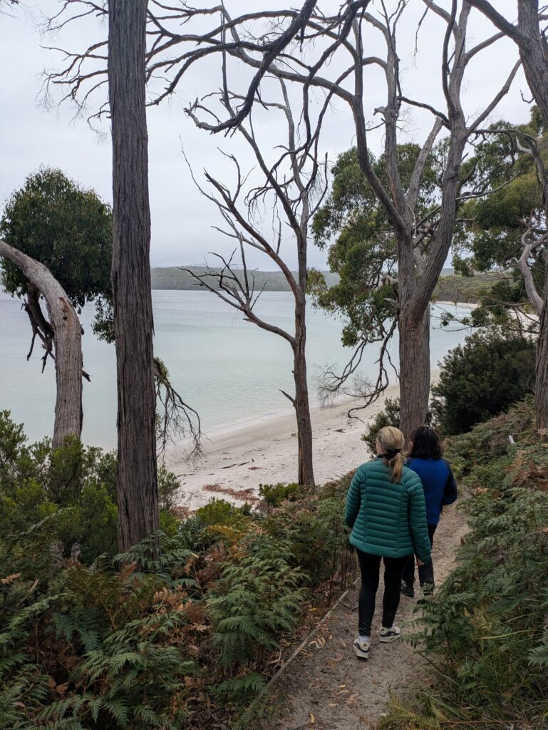 2 women walking down a path to a deserted white sandy beach on a grey, cold day