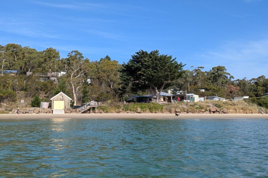 A view from the water, looking back to a shack and boathouse, with a man and dog walking along the beach in front
