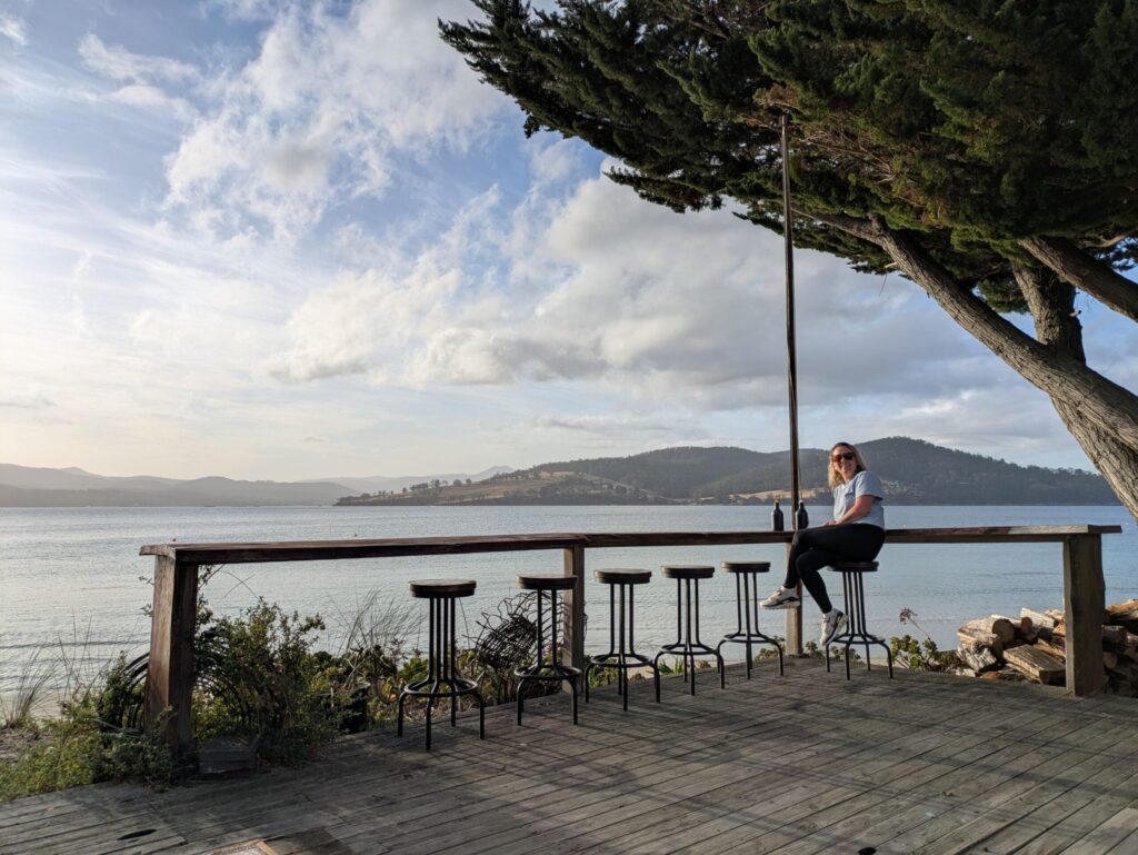 A woman sitting on a high stool at an outdoor bar with the ocean and mountains behind her, and the sun slowly setting