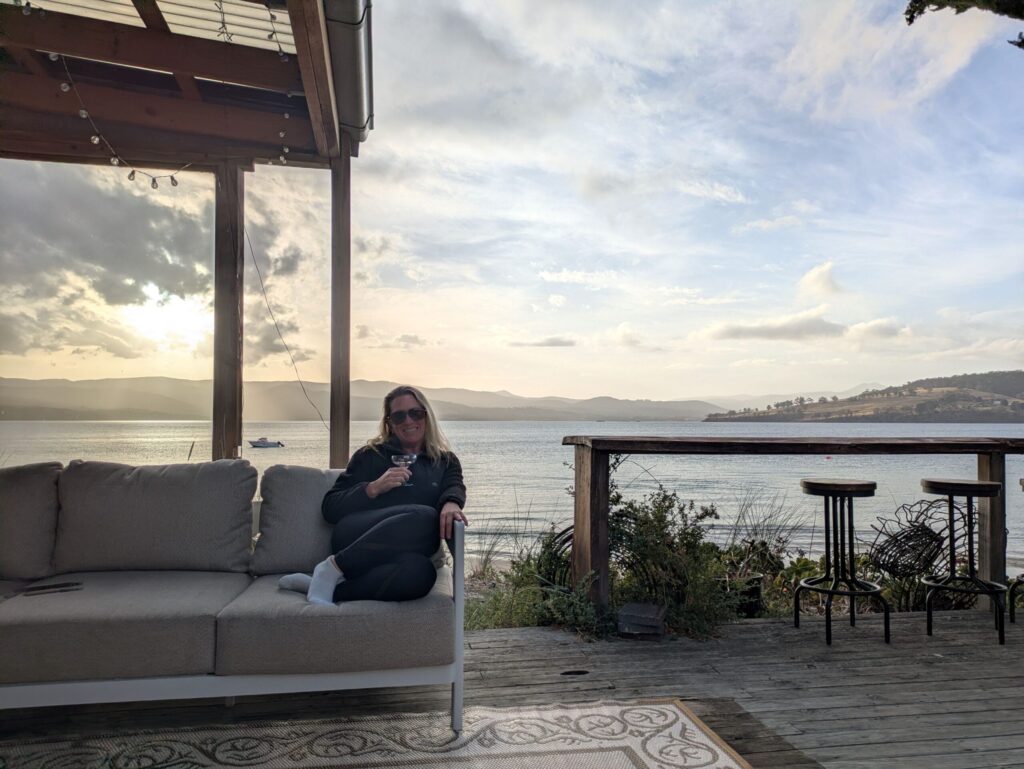 A woman sitting on a comfortable outdoor lounge with the ocean and mountains behind her, and the sun slowly setting