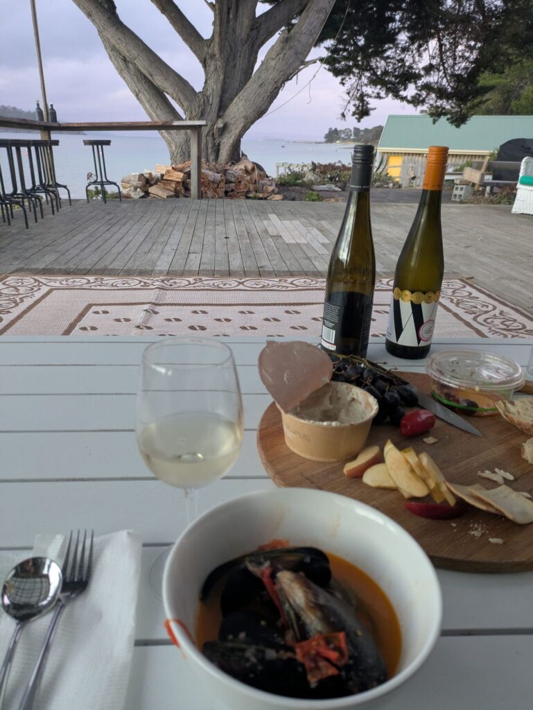 A bowl of mussels, a cheese board and 2 bottles of wine on a table with a large tree and the ocean in the background