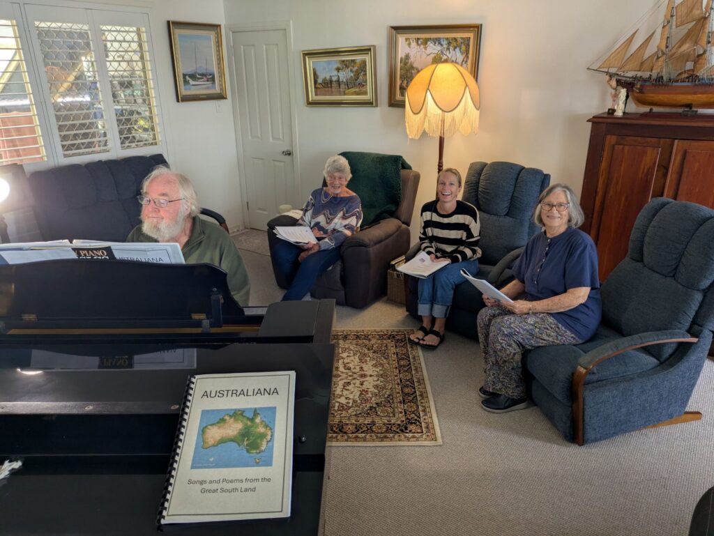 Three women seated in comfy couches, singing along to a man playing a grand piano in a lounge room