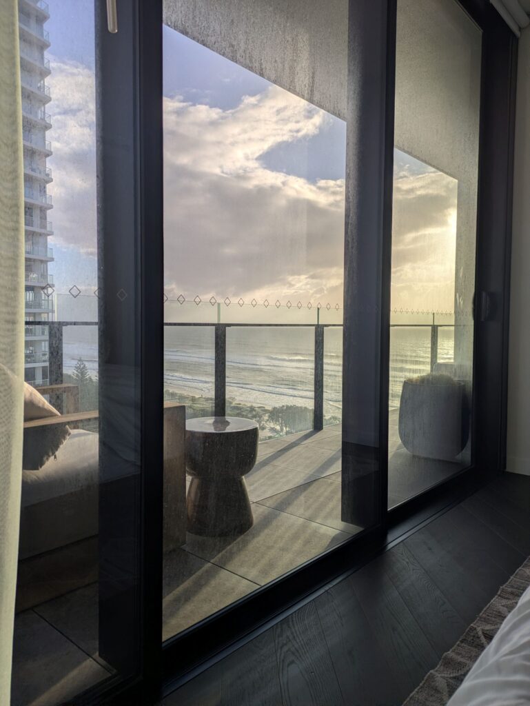 Looking through tall glass windows across a balcony to the ocean as the sun rises