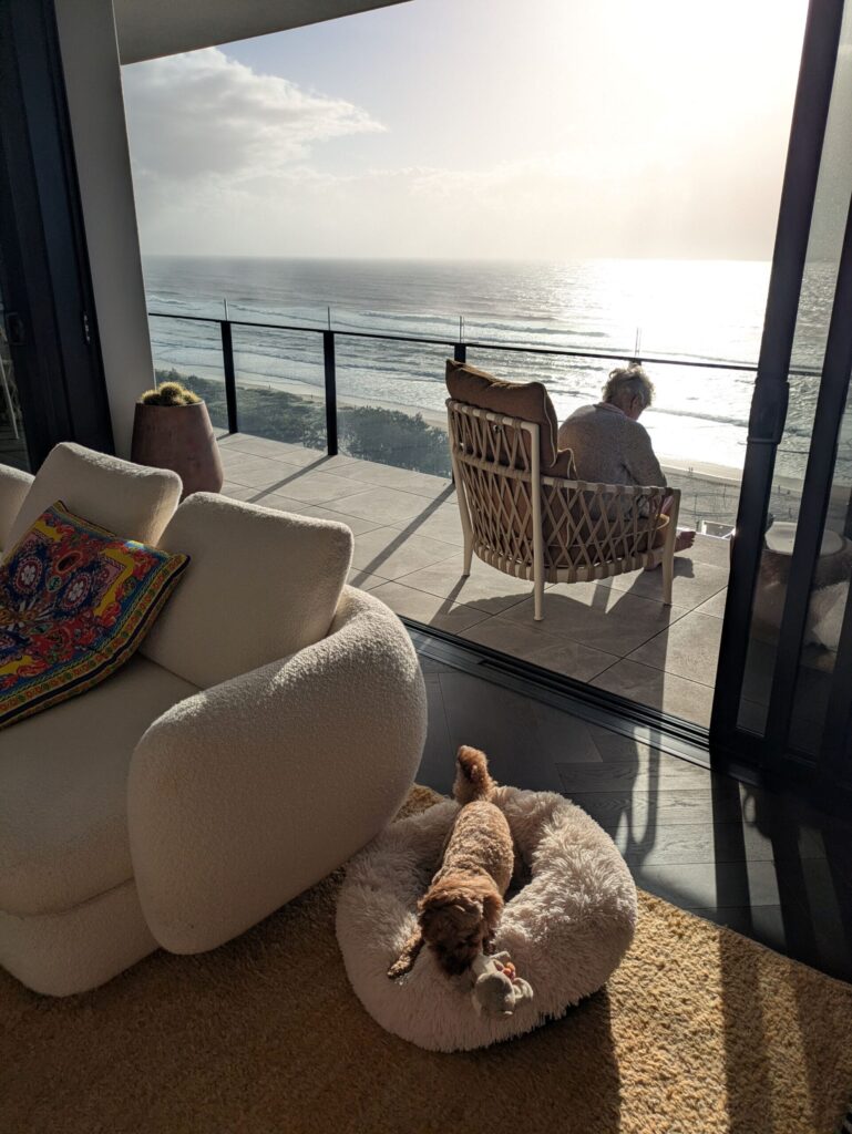 A woman in the distance sitting on a balcony overlooking the ocean. A dog in the foreground sleeping in a bean bag