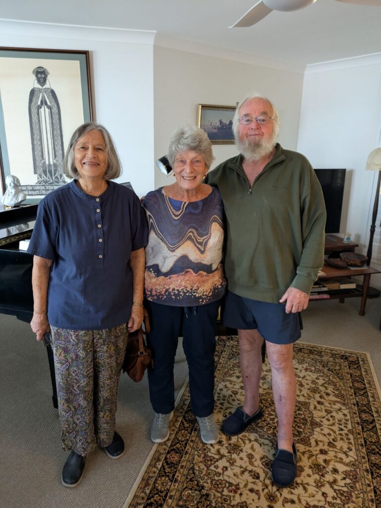 Three friends aged in their 80s, standing together by a piano in a sitting room smiling at the camera