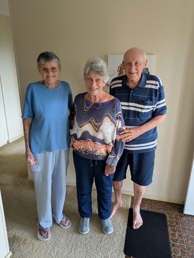 Three friends aged in their 80s, standing together in a hallway smiling at the camera